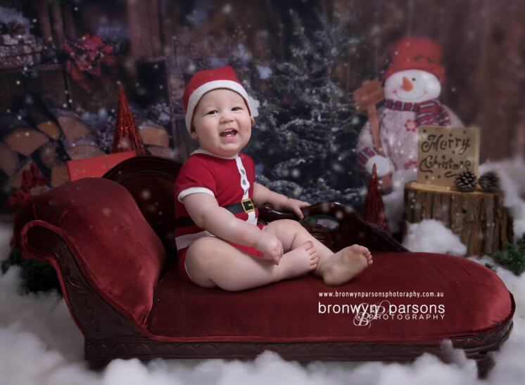 Baby Photography Canberra Smiling Baby Boy in Santa Outfit sitting on red lounge in front of Christmas Tree in the snow